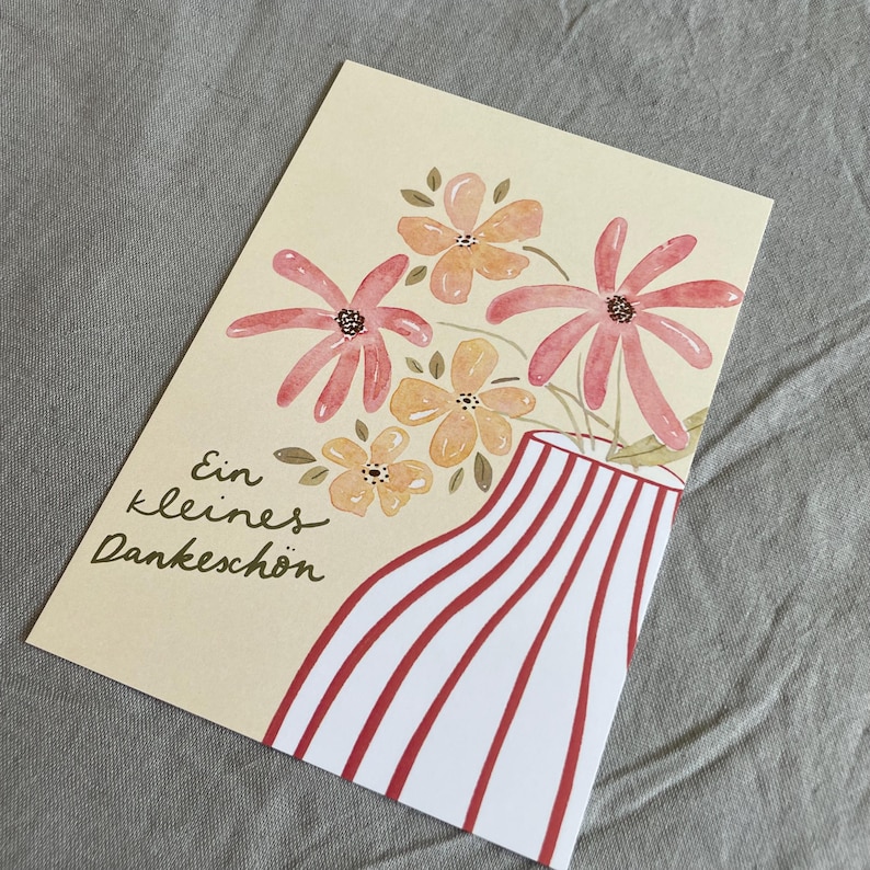 May include: A greeting card featuring a watercolour illustration of flowers in a vase. The vase has red and white stripes. The flowers are pink and orange. The card includes the text "Ein kleines Dankesch&ouml;n".