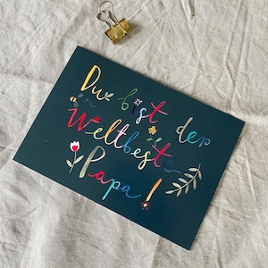 May include: A dark green card with the text "Du bist der weltbeste Papa!" written in rainbow colors. The card is decorated with small flowers and leaves.
