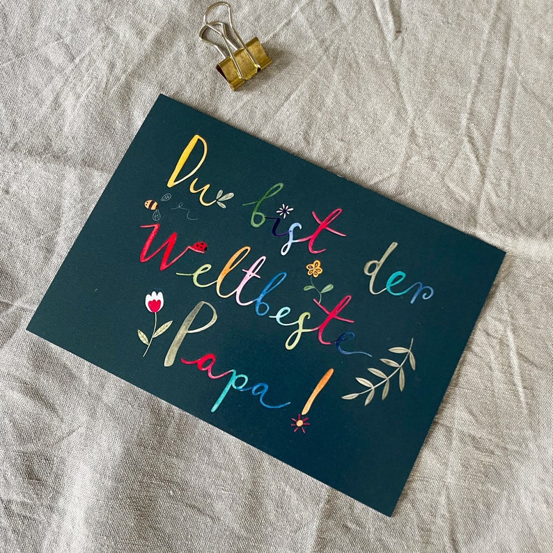 May include: A dark green card with the text "Du bist der weltbeste Papa!" written in rainbow colors. The card is decorated with small flowers and leaves.