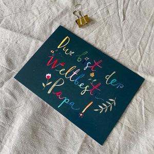 May include: A dark green card with the text "Du bist den weltbeste Papa!" written in colorful letters. The letters are decorated with small flowers, bees, and leaves.