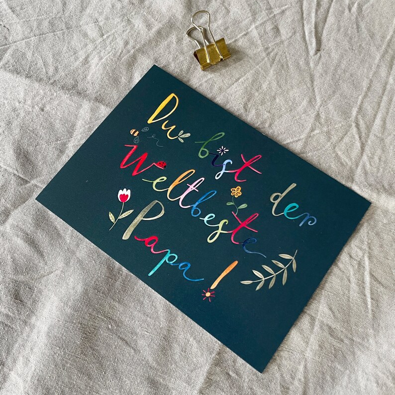 May include: A dark green card with the text "Du bist den weltbeste Papa!" written in colorful letters. The letters are decorated with small flowers, bees, and leaves.