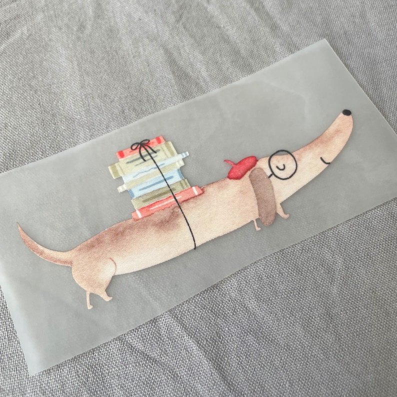 May include: A brown dachshund with a red beret and a stack of books on its back. The dachshund is looking to the right. The background is a light gray.