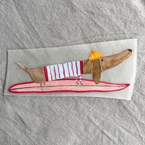 May include: A brown and white dachshund wearing a yellow hat and red and white striped shirt is riding a pink surfboard.