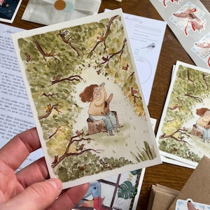Op de afbeelding: Een hand die een aquarelprint vasthoudt van een persoon en een hond in een bos. De print is omgeven door andere briefpapierartikelen, waaronder stickers van vogels en teckels, en een kleine envelop met een ronde sticker.