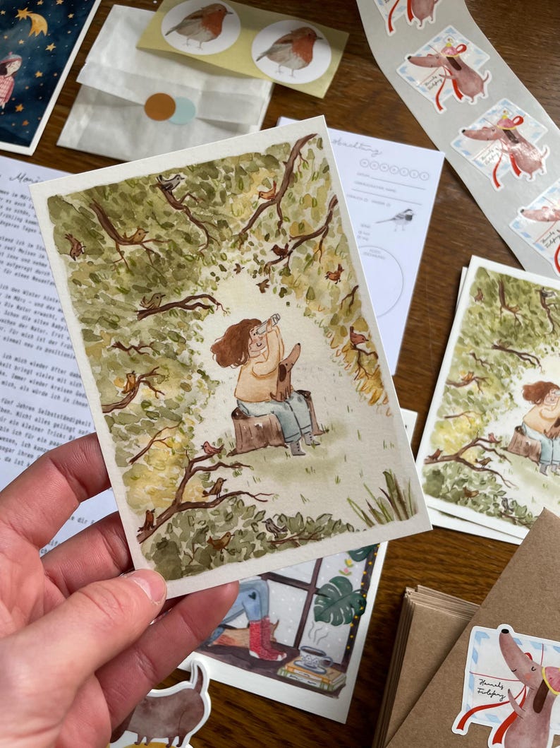 Op de afbeelding: Een hand die een aquarelprint vasthoudt van een persoon en een hond in een bos. De print is omgeven door andere briefpapierartikelen, waaronder stickers van vogels en teckels, en een kleine envelop met een ronde sticker.