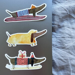 May include: Three dachshund dog stickers with different winter activities. The first sticker shows a dachshund wearing a blue and white striped shirt and a yellow hat, riding a surfboard. The second sticker shows a dachshund wearing a yellow raincoat and a red hat, walking on a white background. The third sticker shows a dachshund wearing a red and white sweater and a red hat, skiing on a white background.