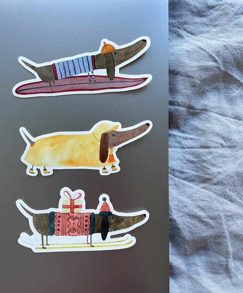 May include: Three dachshund dog stickers with different winter activities. The first sticker shows a dachshund wearing a blue and white striped shirt and a yellow hat, riding a surfboard. The second sticker shows a dachshund wearing a yellow raincoat and a red hat, walking on a white background. The third sticker shows a dachshund wearing a red and white sweater and a red hat, skiing on a white background.