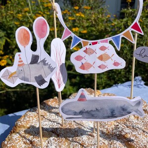 May include: A cake decorated with a whale, a fish, and a seagull, all wearing party hats. The cake is also decorated with a string of colorful flags and a cloud with fish.