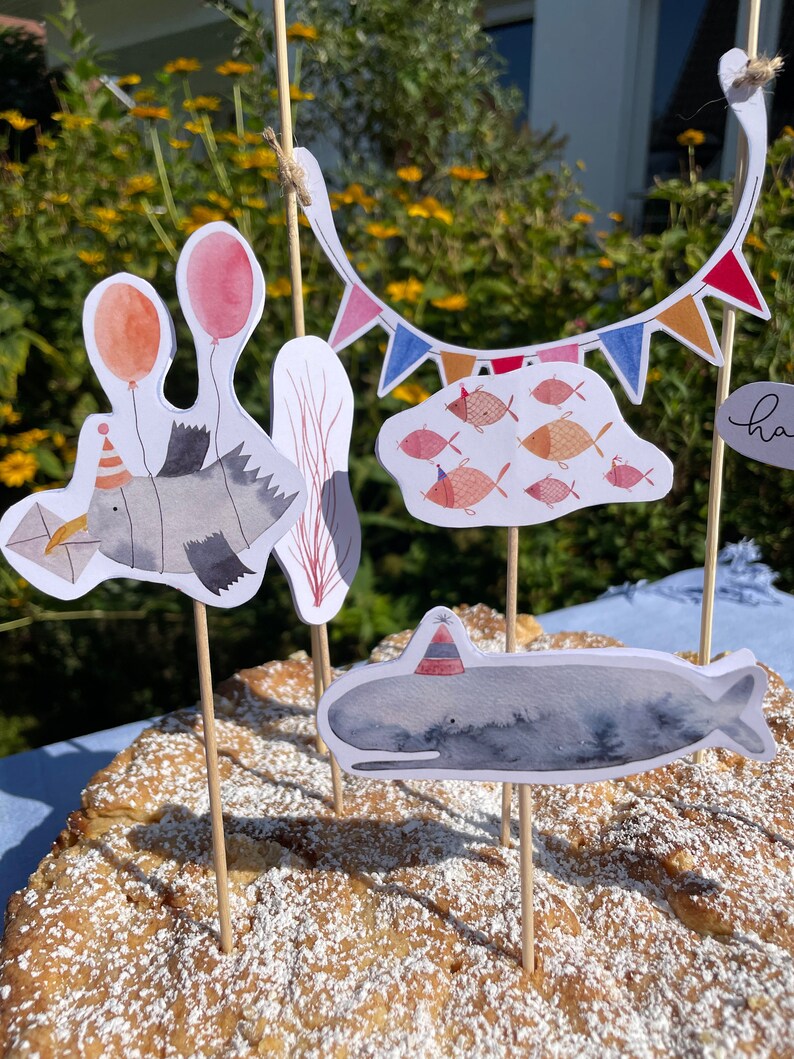 May include: A cake decorated with a whale, a fish, and a seagull, all wearing party hats. The cake is also decorated with a string of colorful flags and a cloud with fish.