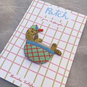 May include: Embroidered patch of a cartoon turtle in a baseball cap, relaxing in a green and red checkered basket. The patch is on a card with the word "Patch" and the text "zum Aufb&uuml;geln".