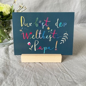 May include: A dark blue card with the text "Du bist der weltbeste Papa!" in colorful lettering. The card is decorated with small flowers and leaves. The card is standing on a wooden easel.