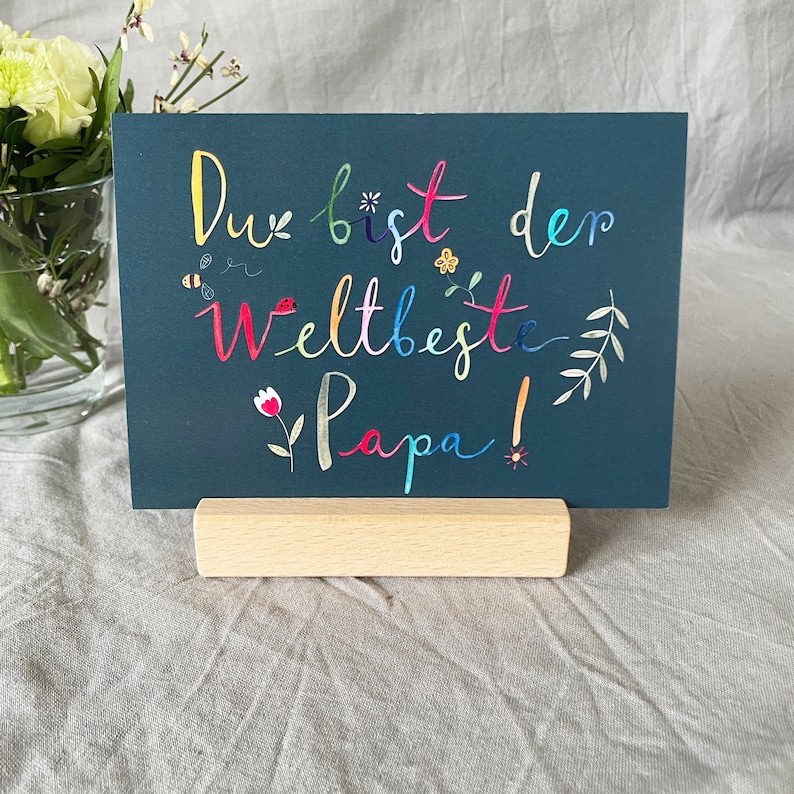 May include: A dark blue card with the text "Du bist der weltbeste Papa!" in colorful lettering. The card is decorated with small flowers and leaves. The card is standing on a wooden easel.