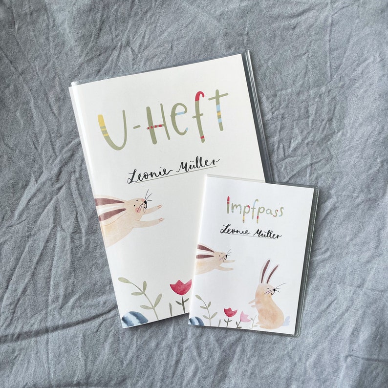 May include: Two white paper notebooks with colorful illustrations of bunnies and flowers. The larger notebook says "U-Heft" and "Leonie M&uuml;ller". The smaller notebook says "Impfpass" and "Leonie M&uuml;ller".