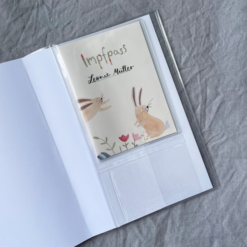 May include: A white booklet with a clear plastic cover. The cover has a colorful illustration of two rabbits and the text "Impfpass Leonie M&uuml;ller".
