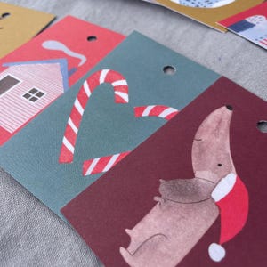K&ouml;nnte beinhalten: Nahaufnahme von weihnachtlichen Geschenkanh&auml;ngern. Ein Anh&auml;nger zeigt ein rotes Haus mit blauen Akzenten, ein anderer ein Zuckerstangen-Design und ein dritter einen Dackel mit Weihnachtsm&uuml;tze. Jeder Anh&auml;nger hat ein Loch zum Befestigen.