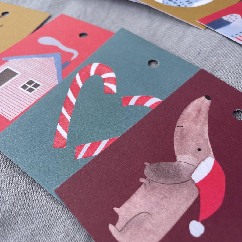 K&ouml;nnte beinhalten: Nahaufnahme von weihnachtlichen Geschenkanh&auml;ngern. Ein Anh&auml;nger zeigt ein rotes Haus mit blauen Akzenten, ein anderer ein Zuckerstangen-Design und ein dritter einen Dackel mit Weihnachtsm&uuml;tze. Jeder Anh&auml;nger hat ein Loch zum Befestigen.