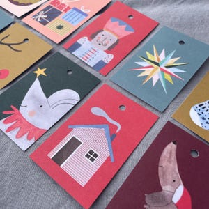 K&ouml;nnte beinhalten: Eine Sammlung rechteckiger Geschenkanh&auml;nger mit verschiedenen festlichen Illustrationen. Designs umfassen einen Nussknacker, einen Stern, ein Haus und einen Hund. Jeder Anh&auml;nger hat oben ein Loch zum Auff&auml;deln. Die Anh&auml;nger sind in Rot-, Gr&uuml;n-, Blau- und Goldt&ouml;nen gehalten.