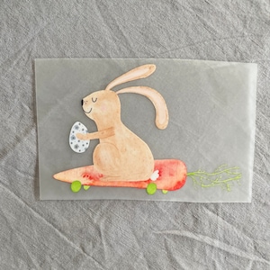 May include: A brown bunny rabbit with a white egg in its paw is riding a red carrot skateboard with green wheels. The bunny is smiling and has a white tail.