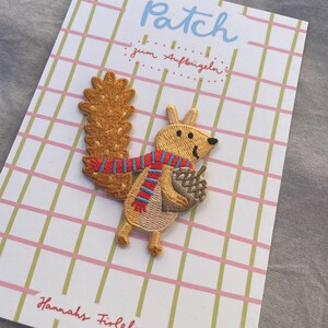May include: Embroidered squirrel patch with a red and blue scarf, holding an acorn. The patch is on a card with the word "Patch" in light blue and the text "zum Aufb&uuml;geln" in red. The background has a pink and green grid pattern.