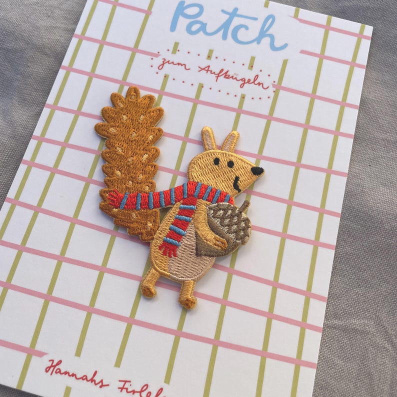 May include: Embroidered squirrel patch with a red and blue scarf, holding an acorn. The patch is on a card with the word "Patch" in light blue and the text "zum Aufb&uuml;geln" in red. The background has a pink and green grid pattern.