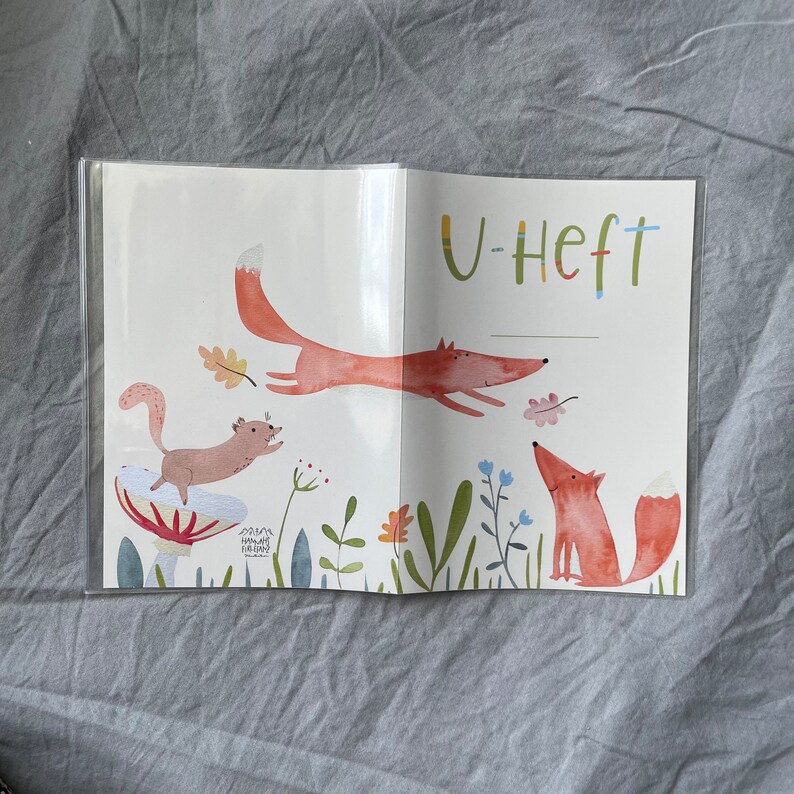 Może przedstawiać: Otwarty notatnik z fantazyjną ilustracją lisa, wiewi&oacute;rki i liści. Lis jest przedstawiony w trakcie skoku, z tekstem "U-Heft" na g&oacute;rze. Grafika ma kolorowy, akwarelowy styl z zabawnym motywem inspirowanym naturą.