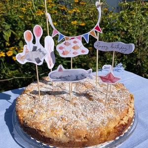 May include: A round cake with a light brown crust and a white powdered sugar topping. The cake is decorated with colorful paper cutouts of fish, a whale, a bird, balloons, and a banner that reads "happy birthday!"