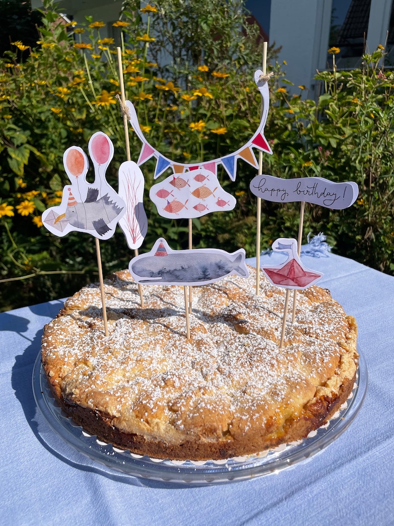 May include: A round cake with a light brown crust and a white powdered sugar topping. The cake is decorated with colorful paper cutouts of fish, a whale, a bird, balloons, and a banner that reads "happy birthday!"