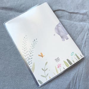 May include: A white notebook with a clear plastic cover. The cover features a watercolour illustration of a grey elephant, green leaves, and a small orange butterfly. The text "Hannah Firman" is printed in black at the bottom of the cover.
