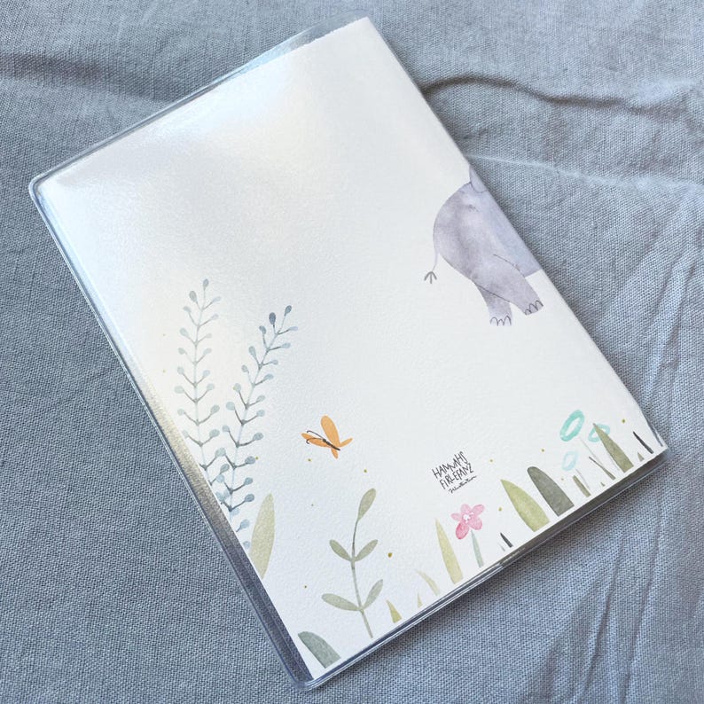 May include: A white notebook with a clear plastic cover. The cover features a watercolour illustration of a grey elephant, green leaves, and a small orange butterfly. The text "Hannah Firman" is printed in black at the bottom of the cover.