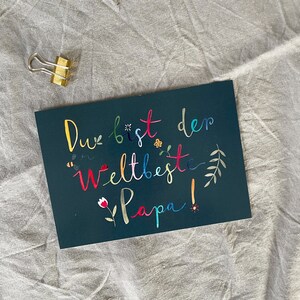 May include: A dark blue card with the text "Du bist der weltbeste Papa!" in colorful script. The text is surrounded by small flowers, bees, and leaves.