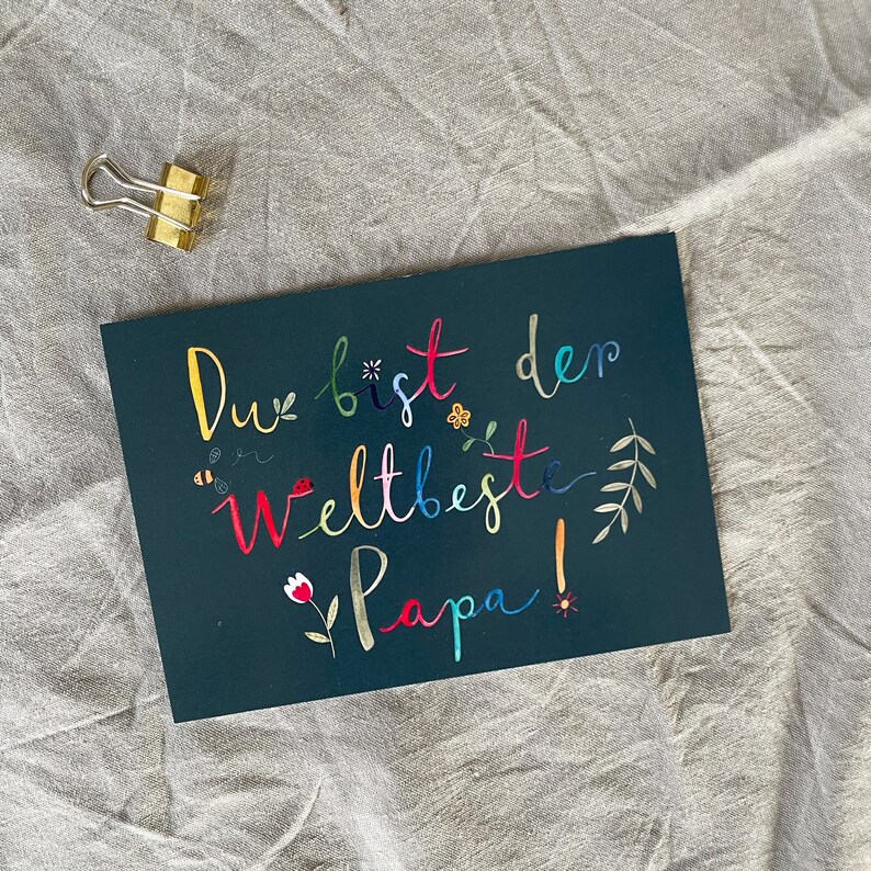May include: A dark blue card with the text "Du bist der weltbeste Papa!" in colorful script. The text is surrounded by small flowers, bees, and leaves.