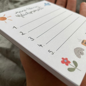 Op de afbeelding: Een witte notitieblok met de handgeschreven tekst "Meine kleinen Gl&uuml;cksmomente" en genummerde lijnen. Het notitieblok bevat kleine illustraties van een zon, een wolk, een bloem en een hond. Het notitieblok wordt in een hand gehouden.