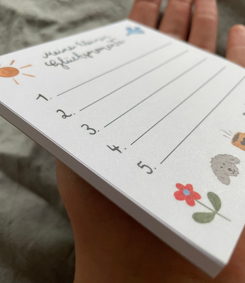 Op de afbeelding: Een witte notitieblok met de handgeschreven tekst "Meine kleinen Gl&uuml;cksmomente" en genummerde lijnen. Het notitieblok bevat kleine illustraties van een zon, een wolk, een bloem en een hond. Het notitieblok wordt in een hand gehouden.