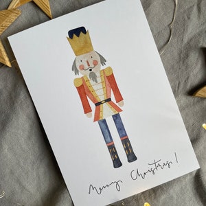 May include: A watercolor illustration of a nutcracker with a red and gold uniform, a black hat, and a gold crown. The nutcracker is standing on a white background with the text "Merry Christmas!" written below.