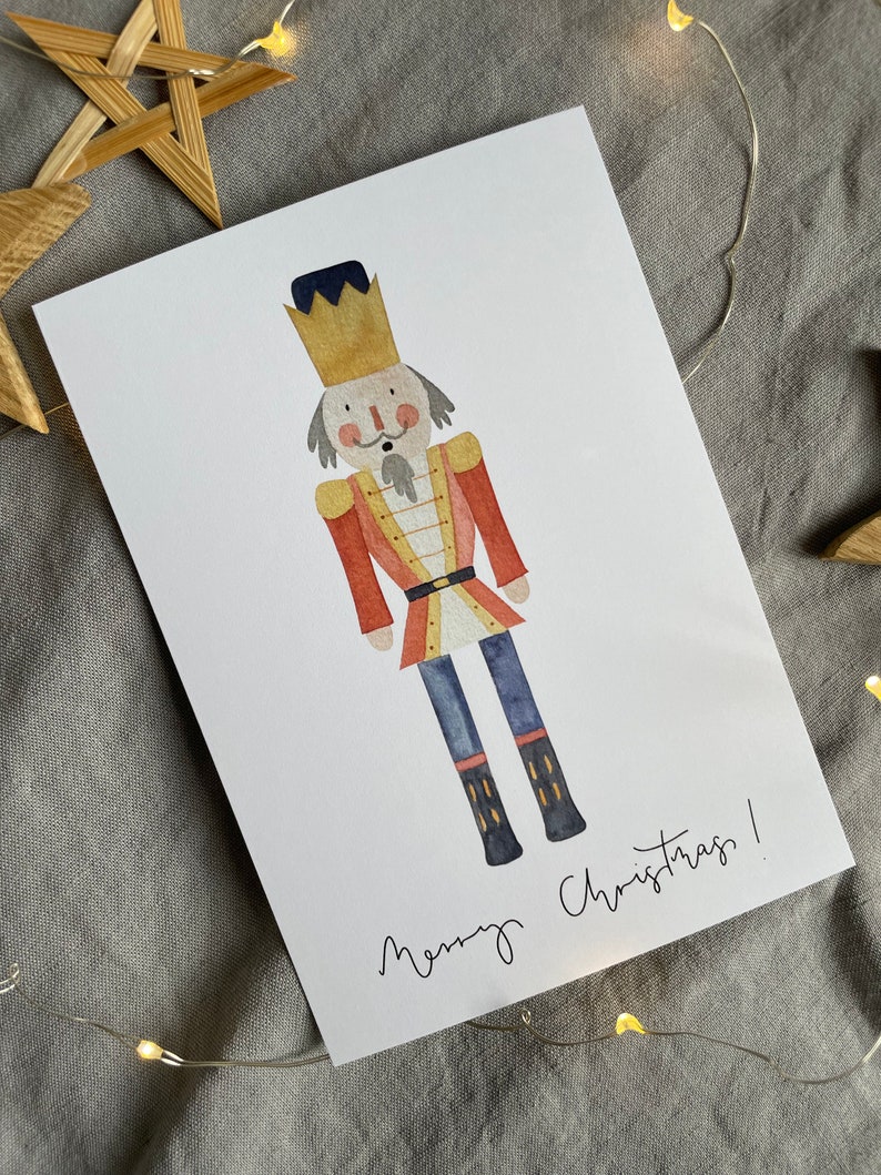 May include: A watercolor illustration of a nutcracker with a red and gold uniform, a black hat, and a gold crown. The nutcracker is standing on a white background with the text "Merry Christmas!" written below.