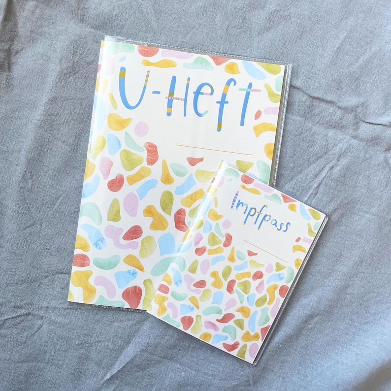 May include: Two clear plastic covered notebooks with colorful abstract patterns. The larger notebook has the text "U-Heft" on the front. The smaller notebook has the text "Impfpass" on the front.