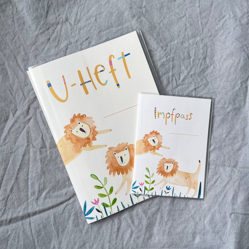 K&ouml;nnte beinhalten: Ein wei&szlig;es Notizbuch mit dem Text "U-Heft" in Gelb und einer bunten Illustration von L&ouml;wen und Pflanzen. Ein kleineres wei&szlig;es Notizbuch mit dem Text "Impfpass" in Schwarz und einer &auml;hnlichen Illustration von L&ouml;wen liegt auf dem gr&ouml;&szlig;eren Notizbuch.
