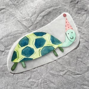 Puede incluir: Una tortuga verde y azul con un gorro de fiesta rosa y blanco. La tortuga tiene una cara feliz y sonríe.