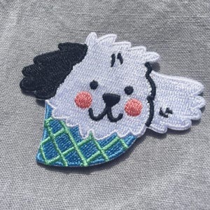 May include: An embroidered patch featuring a white dog head with one black ear, two black eyes, a black nose, and pink cheeks. The dog wears a blue and green plaid bandana. The patch has a white border and is stitched with detailed thread work.