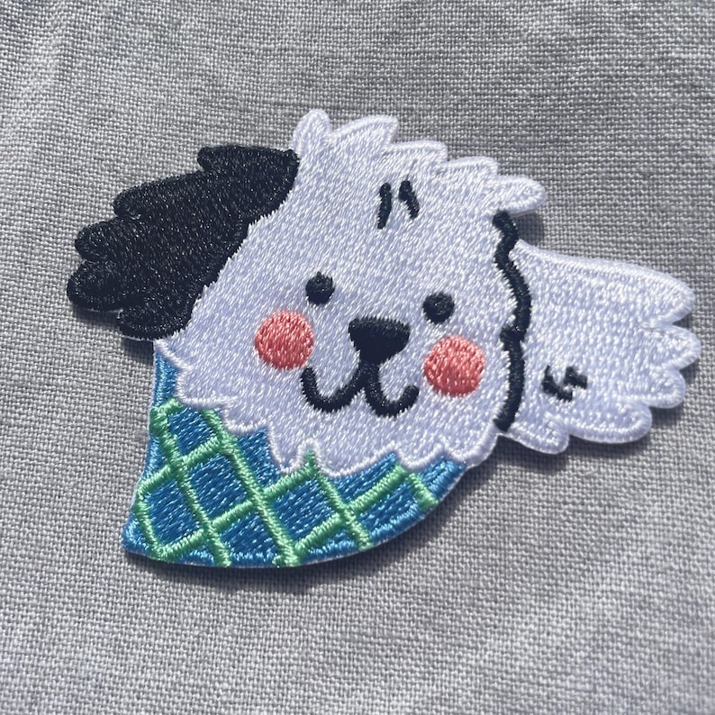 May include: An embroidered patch featuring a white dog head with one black ear, two black eyes, a black nose, and pink cheeks. The dog wears a blue and green plaid bandana. The patch has a white border and is stitched with detailed thread work.