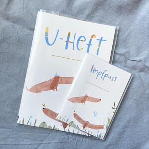 May include: Two white covered notebooks with dachshund illustrations. The larger notebook has the text "U-Heft" in blue and yellow. The smaller notebook has the text "Impfpass" in blue. Both notebooks have a clear plastic cover.