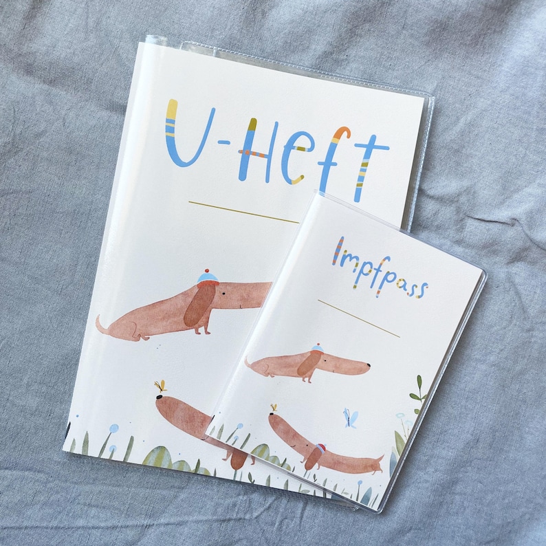 May include: Two white covered notebooks with dachshund illustrations. The larger notebook has the text "U-Heft" in blue and yellow. The smaller notebook has the text "Impfpass" in blue. Both notebooks have a clear plastic cover.
