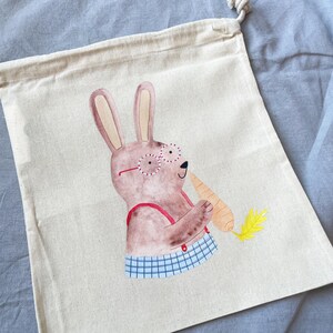 May include: A beige canvas tote bag with a brown bunny wearing red glasses and a blue and white checkered shirt holding a carrot. The bunny is holding the carrot in its right paw and has a happy expression on its face.