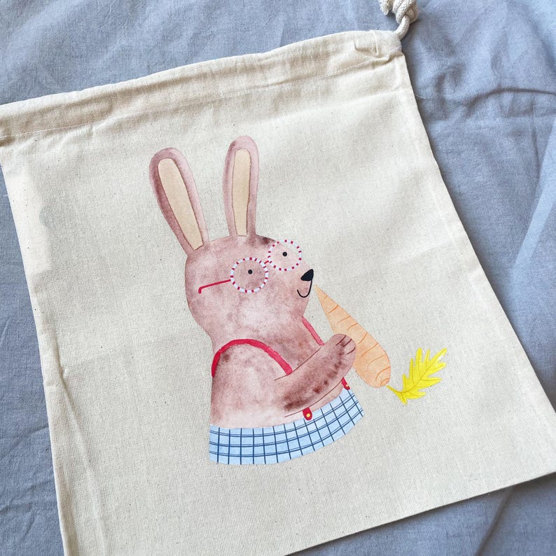 May include: A beige canvas tote bag with a brown bunny wearing red glasses and a blue and white checkered shirt holding a carrot. The bunny is holding the carrot in its right paw and has a happy expression on its face.