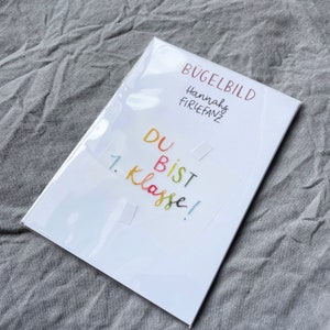 Może przedstawiać: Biała kartka z nadrukowanym tekstem "B&uuml;gelbild Hannahs Firlefanz Du bist 1. Klasse!". Kartka znajduje się w przezroczystej plastikowej osłonie.
