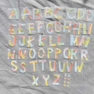 Könnte beinhalten: Eine Sammlung farbenfroher Buchstaben im Aquarellstil, die jeweils einzeln ausgeschnitten und auf einer transparenten Rückseite befestigt sind. Die Buchstaben sind mit pastellfarbenen Streifen, Punkten und anderen Mustern verziert. Die Buchstaben sind auf einer grauen Oberfläche angeordnet.