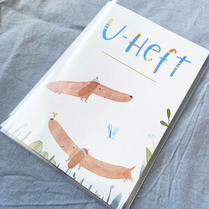 May include: A clear plastic cover encases a notebook with a whimsical illustration. The cover features two dachshunds wearing hats, with the text "U-Heft" in blue and yellow. The background includes butterflies and floral elements.