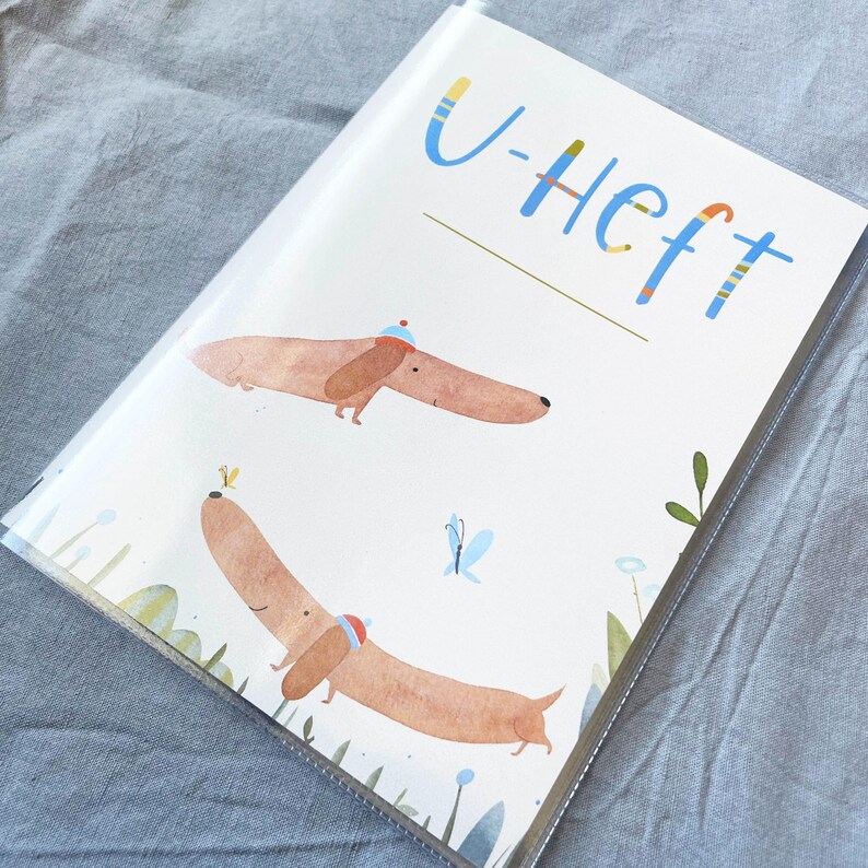 May include: A clear plastic cover encases a notebook with a whimsical illustration. The cover features two dachshunds wearing hats, with the text "U-Heft" in blue and yellow. The background includes butterflies and floral elements.