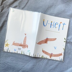 May include: Open notebook with illustrations of dachshunds and the text "U-Heft". The illustrations feature brown dachshunds with hats and butterflies. The notebook is in a clear plastic cover.