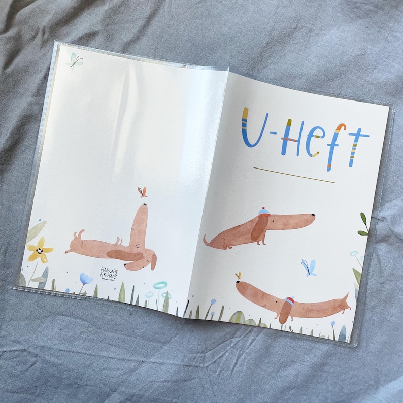May include: Open notebook with illustrations of dachshunds and the text "U-Heft". The illustrations feature brown dachshunds with hats and butterflies. The notebook is in a clear plastic cover.